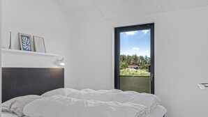 Ein Schlafzimmer mit einem Bett, einem Nachttisch, einer Wandlampe und einem Fenster mit Blick auf Grünflächen und ein Haus.