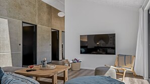 Ein modernes Wohnzimmer mit einer Couch, einem Couchtisch und einem Sessel. An der Wand ist ein Fernseher angebracht und eine Tür führt in einen anderen Raum.