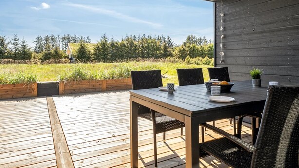 Eine Holzterrasse im Freien mit Tisch und Stühlen, mit Blick auf ein Rasenfeld und Bäume.