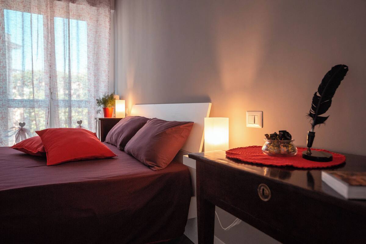 A neatly made bed with red and brown bedding, a bedside table with a lamp, a decorative item, and a window with sheer curtains.