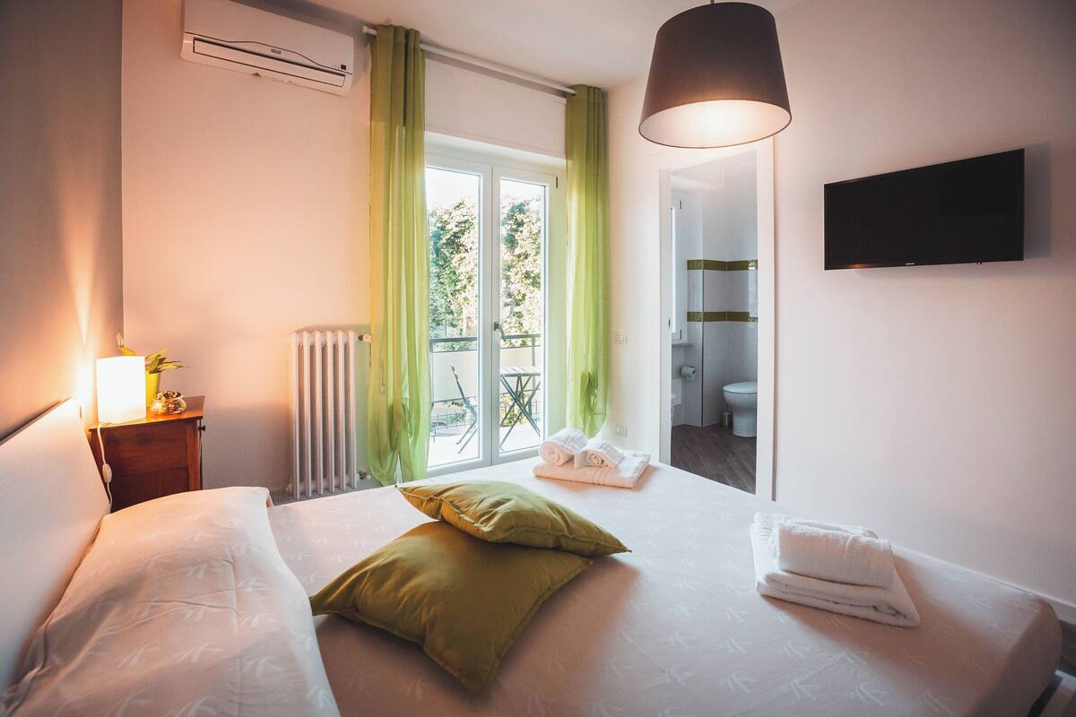 A hotel room with a bed, a bedside lamp, a radiator, a window with curtains, a TV mounted on the wall, and a view of the bathroom.