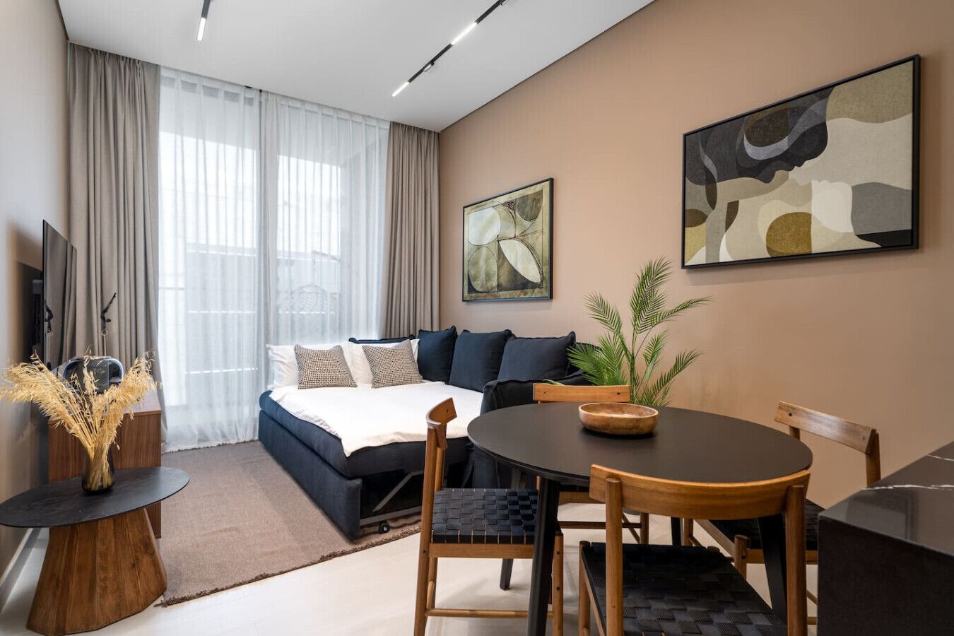 A modern hotel room with a bed, a dining table, and chairs. There is a plant on the table and a TV on the left side.