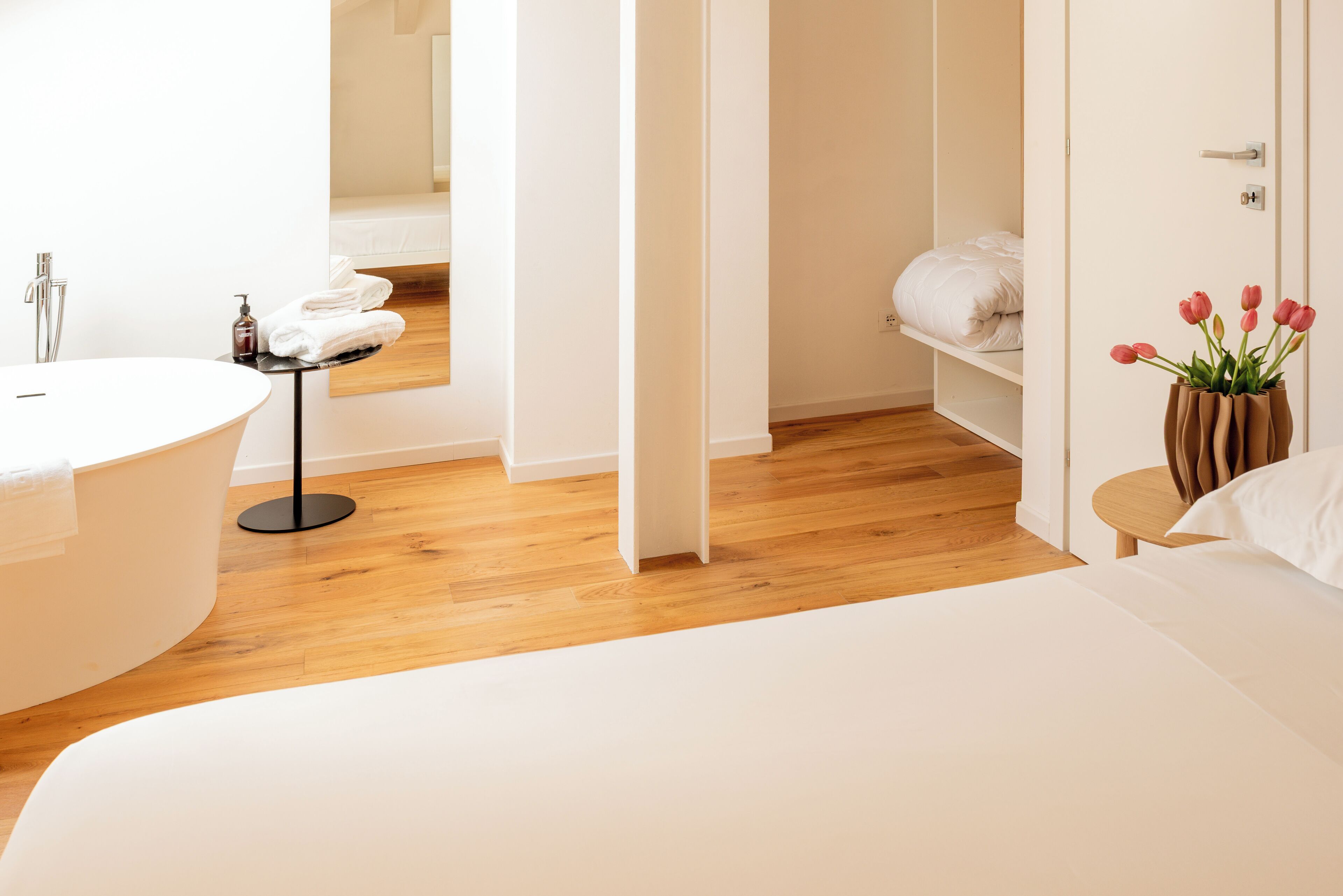 Una camera d'albergo moderna con una vasca da bagno bianca e grande, pavimento in legno, un letto bianco e un tavolino con un vaso di fiori.