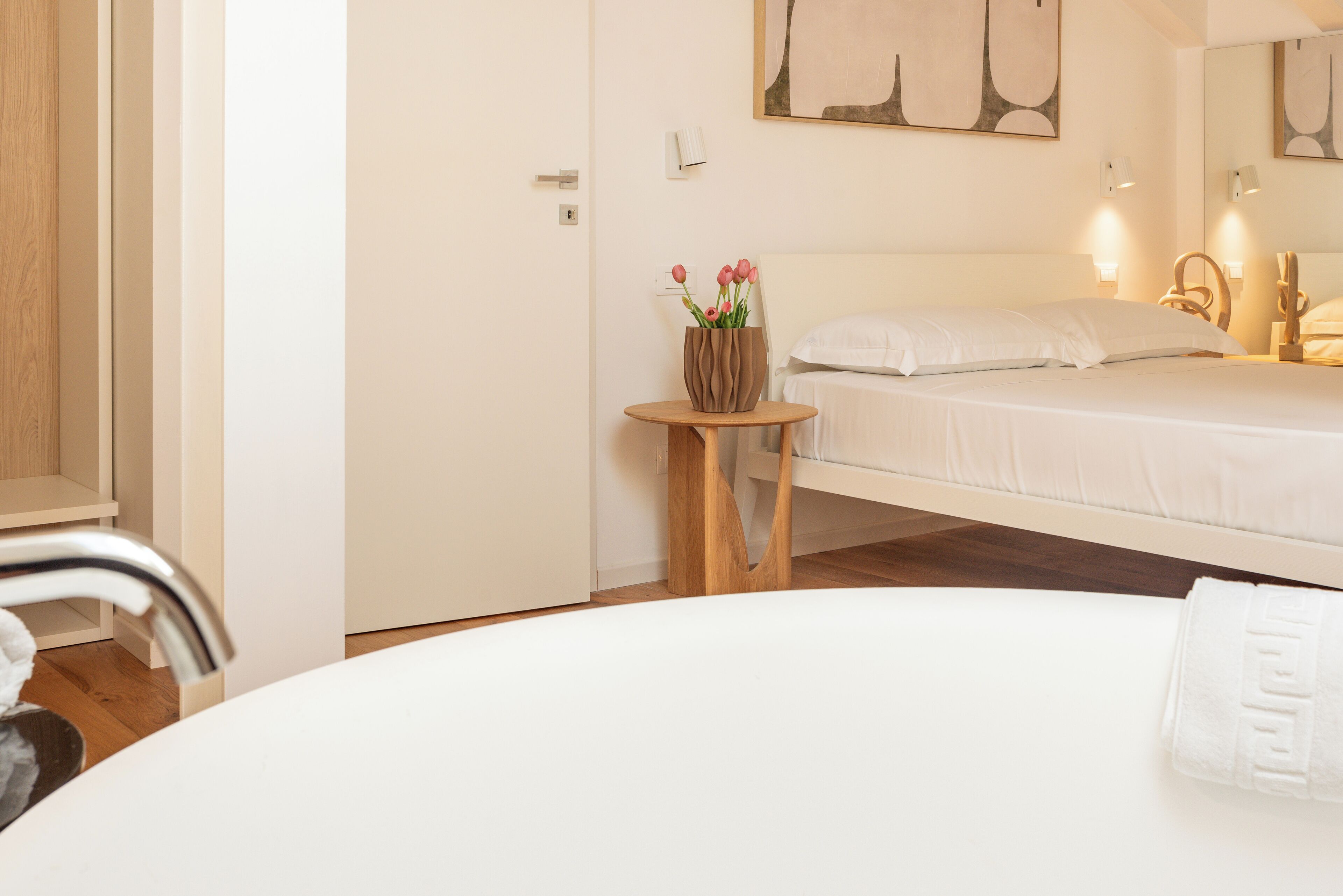 A hotel room with a bed, a small wooden table with a vase of flowers, a round white table, and a white chair.