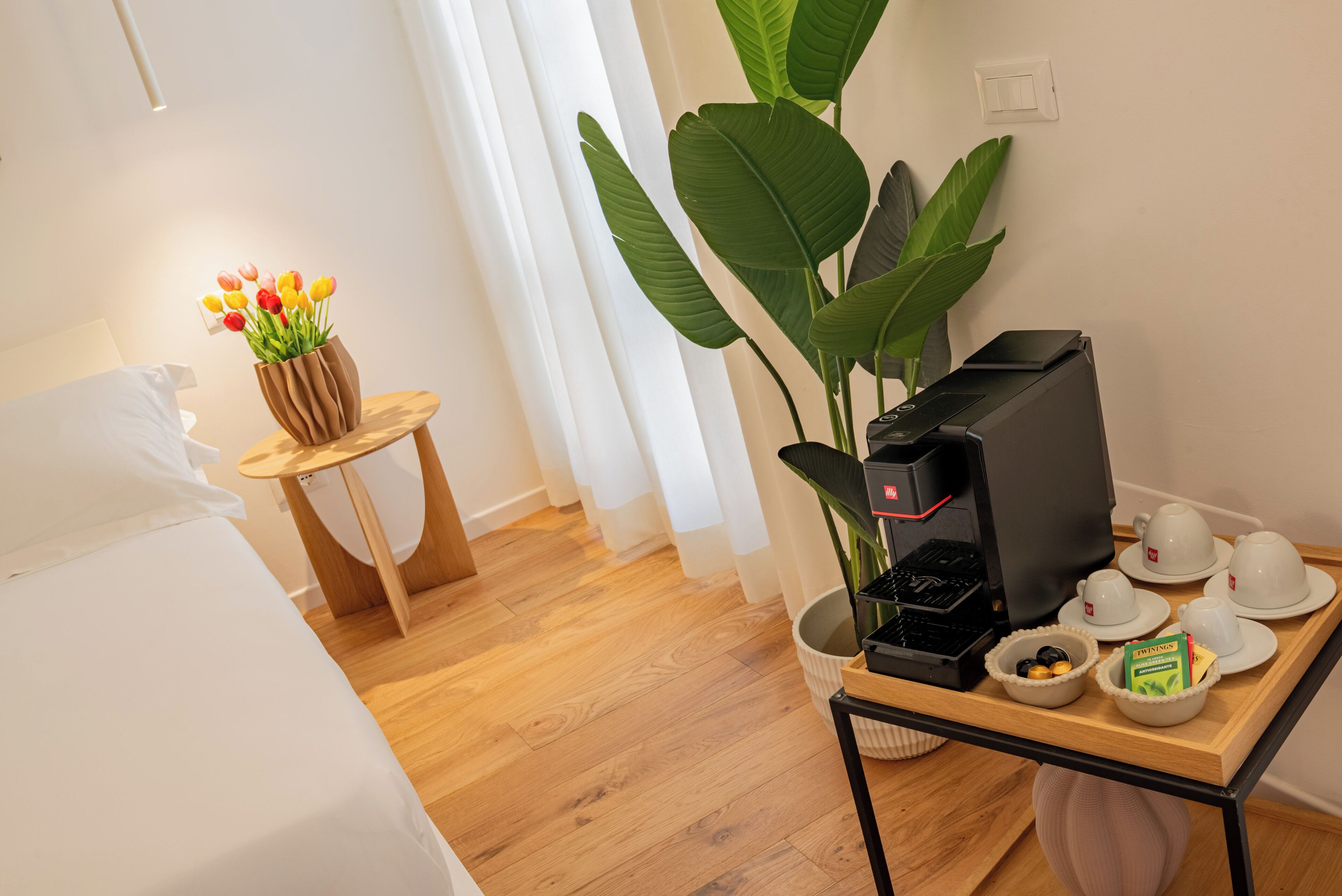A hotel room with a bed, a small wooden table with a coffee machine, cups, and a tray with a box of tissues.
