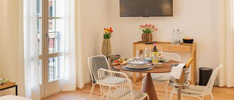 Un comedor con una mesa de madera, sillas blancas, un televisor montado en la pared y un ventanal con cortinas.