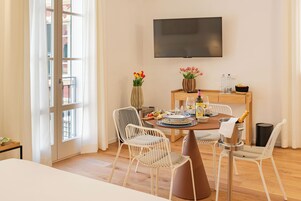Un comedor con una mesa de madera, sillas blancas, un televisor montado en la pared y un ventanal con cortinas.