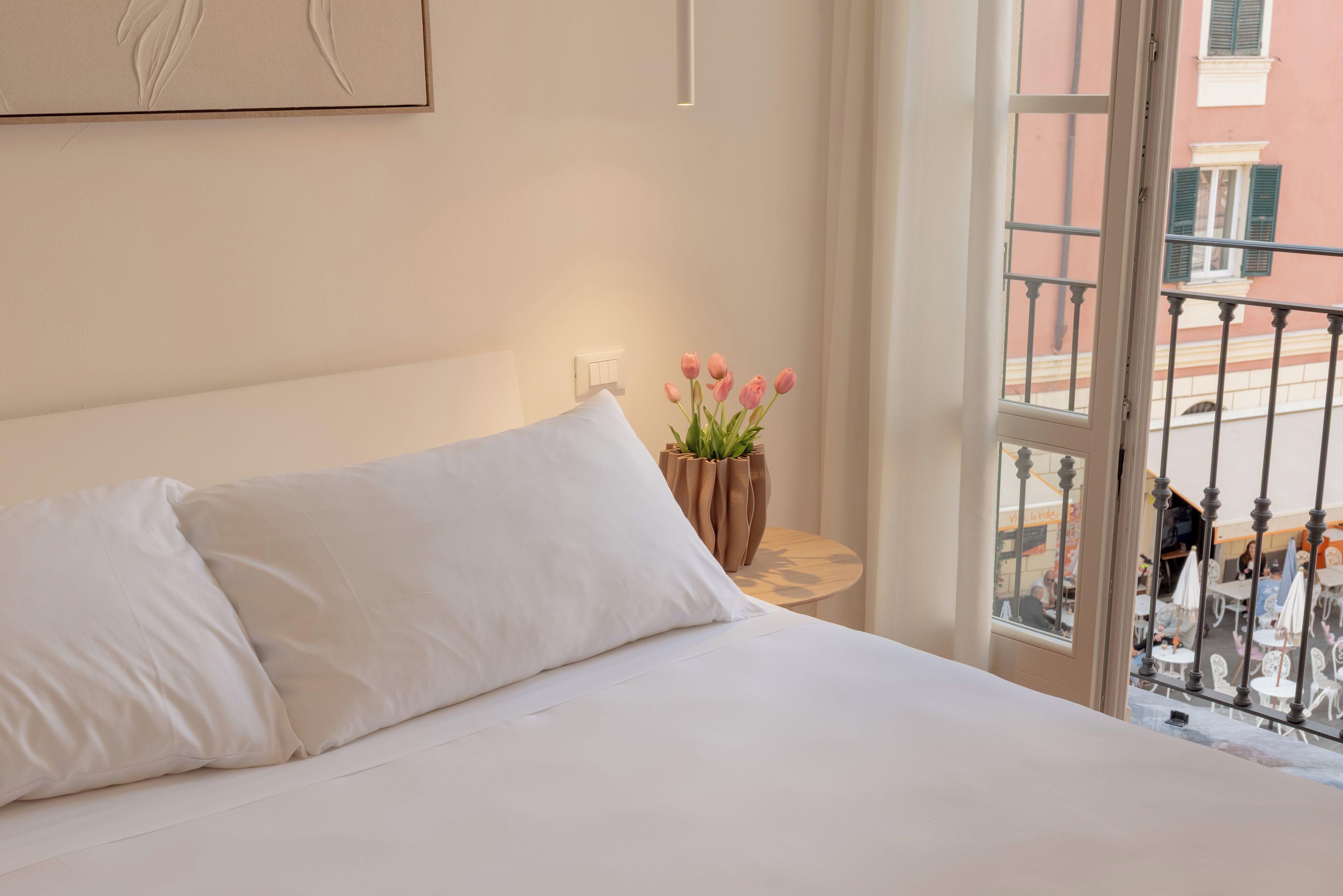 Une chambre d’hôtel avec un lit, des oreillers blancs, un vase de fleurs et une vue sur la rue par la fenêtre.