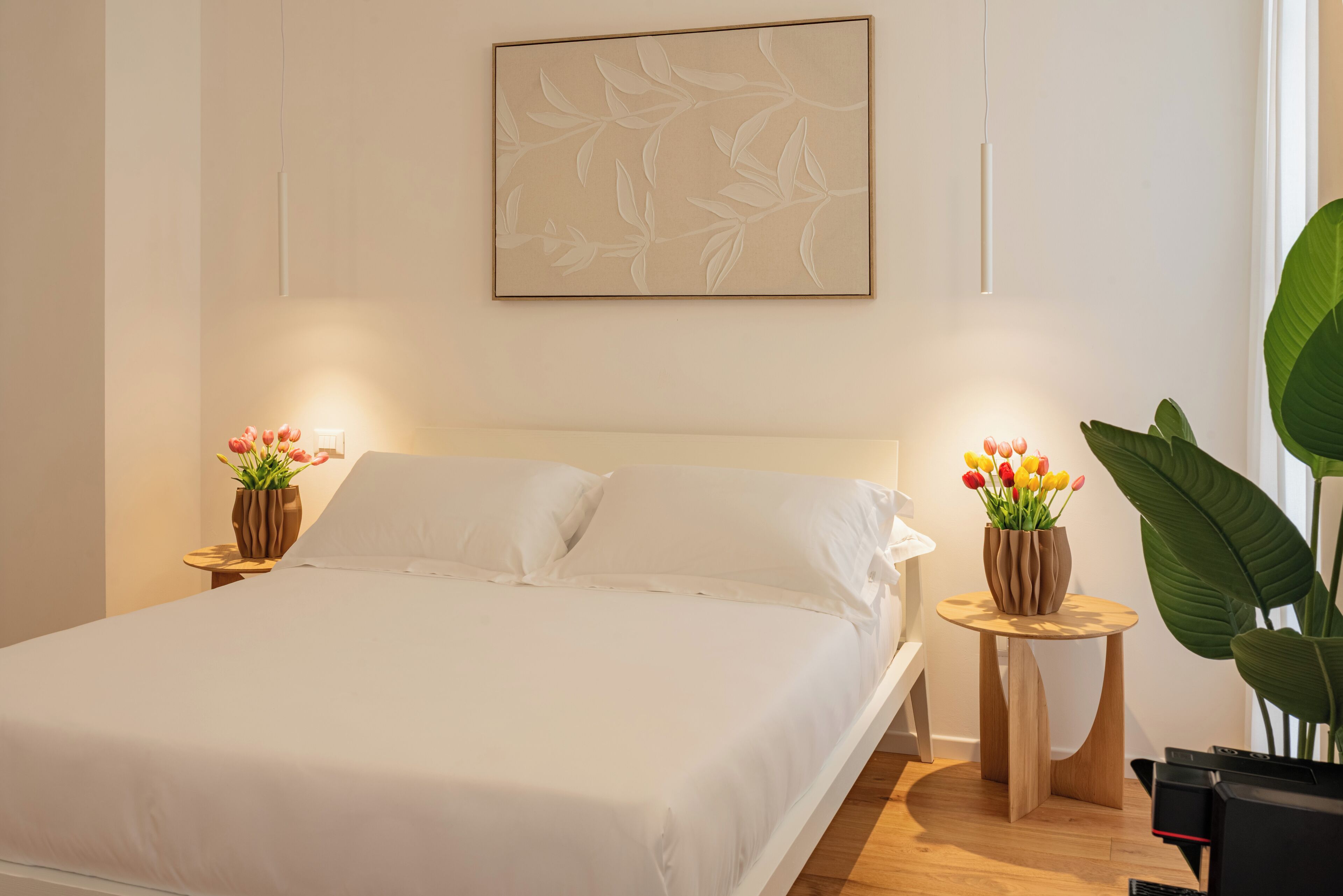 Un dormitorio moderno con una cama grande, dos mesitas de noche con jarrones de flores, una obra de arte en la pared y una planta de gran tamaño.