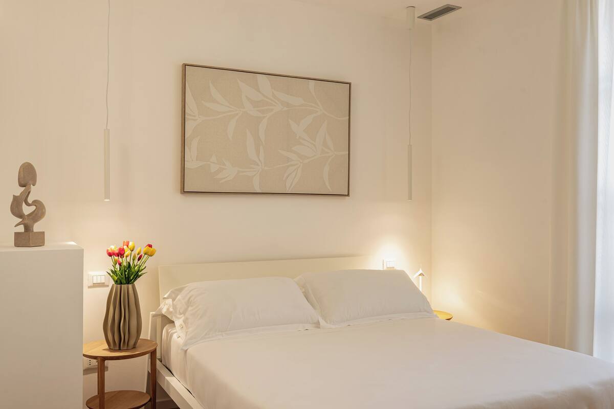 A neatly made bed with white linens, a vase with flowers on a bedside table, and a wall-mounted artwork.
