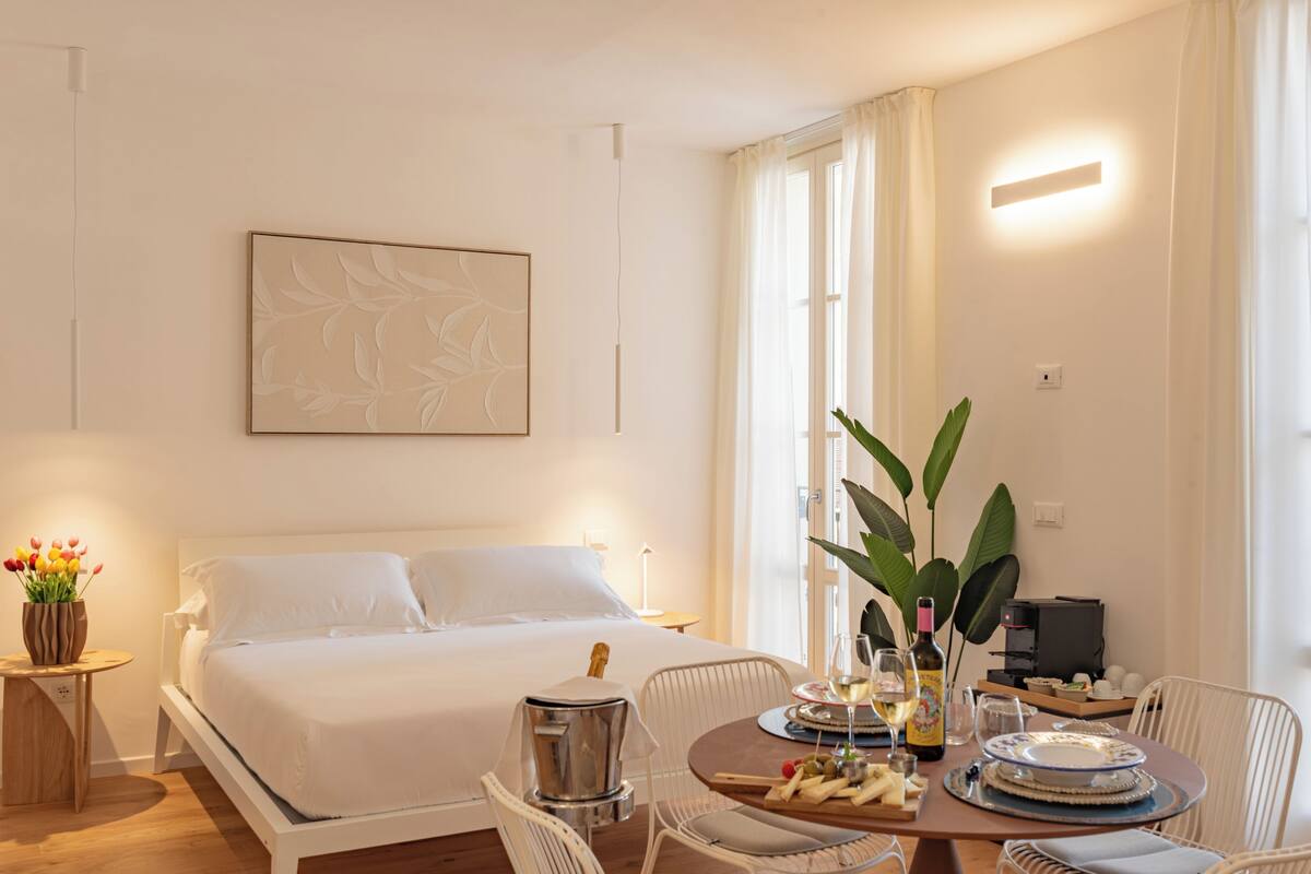 A hotel room with a large bed, a dining table with chairs, a coffee machine, and a large window with curtains.