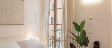 Una habitación de hotel con una cama, un ventanal con cortinas, una lámpara en la pared, una planta y una mesita con una cafetera.