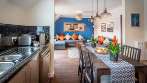 Une cuisine moderne équipée d’une cuisinière intégrée, d’un évier et d’une machine à café. On y trouve également un coin repas avec une table et des chaises, ainsi qu’un canapé agrémenté de coussins orange.