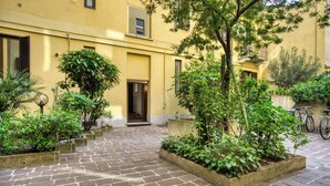 Une cour avec un chemin pavé, entourée de verdure et bordée d’un bâtiment à la façade jaune.