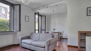 Un salon moderne avec un canapé gris, un plancher en bois, un coin repas comprenant des chaises et une table, et une vue sur le balcon extérieur.