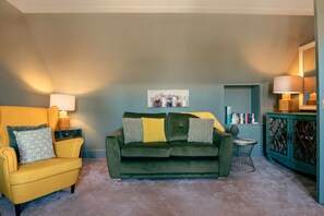 Un salon avec un canapé vert, un fauteuil jaune et une petite table ronde. On y trouve une bibliothèque remplie de livres et d’objets décoratifs.