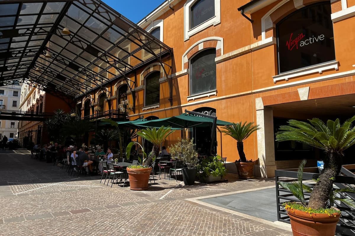 An outdoor café area with tables and chairs, potted plants, and a building with a sign reading "Virgin Active".