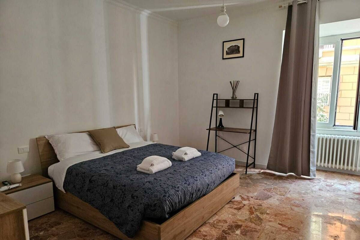 A bedroom with a wooden bed, a nightstand, a bedside table, a radiator, a window with curtains, and a wall-mounted picture.