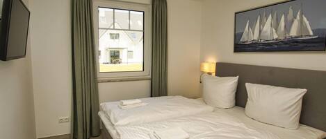 A hotel room with a large bed, a flat-screen TV, a window with curtains, a painting of sailboats, and a lamp.