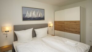 A neatly made bed with white linens, two bedside lamps, a painting of sailboats, and a wooden panel on the wall.