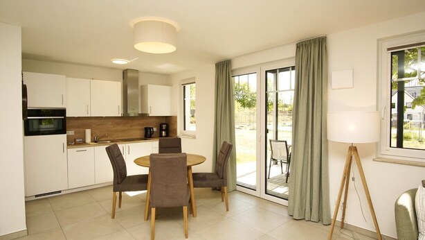 A modern kitchen with white cabinets, a round wooden table, and four chairs. There is a large window with curtains, a floor lamp, and a view of the outdoors.