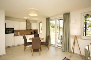 A modern kitchen with white cabinets, a round wooden table, and four chairs. There is a large window with curtains, a floor lamp, and a view of the outdoors. - Beachfront vacation rental with a terrace, access to the pool and sauna - Rex Rugia H-1010 - (Göhren)