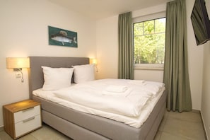 A neatly made bed with white linens and a folded towel, a bedside table with a lamp, and a painting of a fish on the wall.
