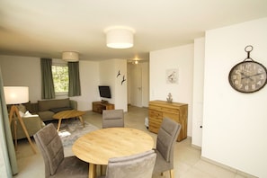 A modern living room with a round wooden table, grey upholstered chairs, a sofa, a TV, and a wooden cabinet. - Beachfront vacation rental with a terrace, access to the pool and sauna - Rex Rugia - Rex (Göhren)