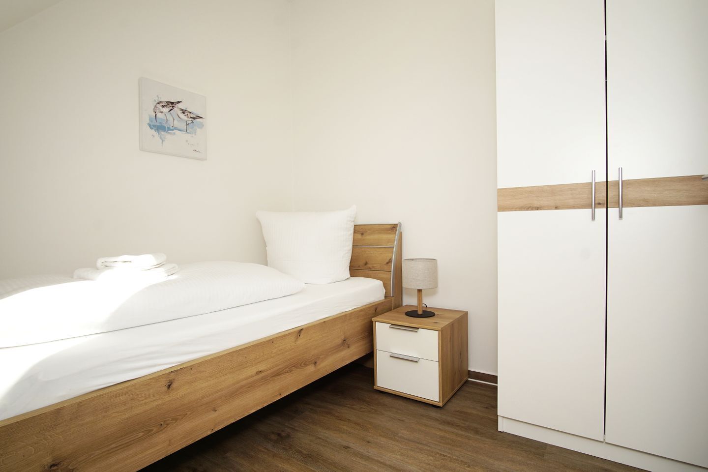 A bedroom with a wooden bed, white bedding, a bedside table with a lamp, and a wardrobe.