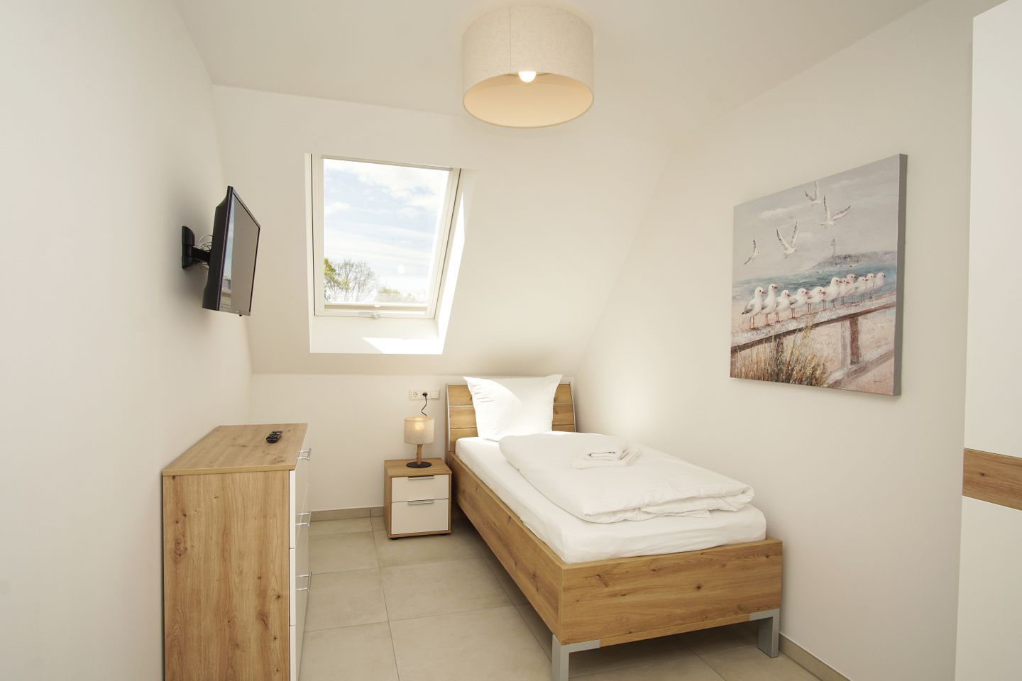 Een slaapkamer met een bed, nachtkastje, lamp, televisie, raam en kunstwerk.