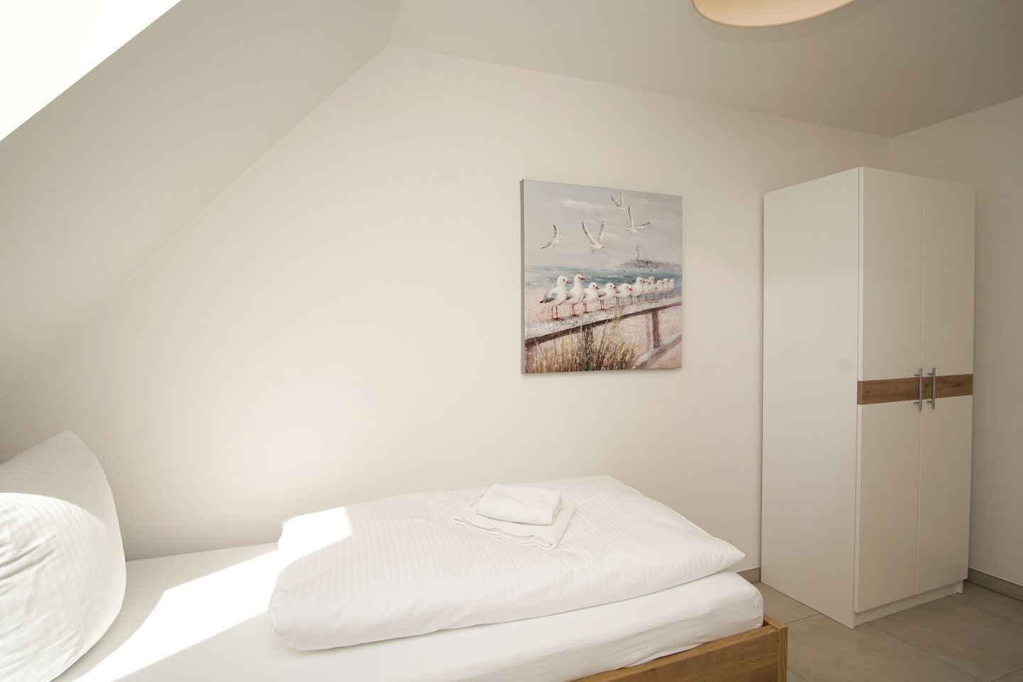 A bedroom with a sloped ceiling, a bed with white bedding, a white wardrobe, and a seagulls painting on the wall.