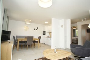 A modern living room with a flat-screen TV, a dining area with a wooden table and chairs, and a kitchen with white cabinets and a built-in oven. - Beachfront vacation rental with a terrace; access to the pool and sauna - Rex Rugia - Rex (Göhren)