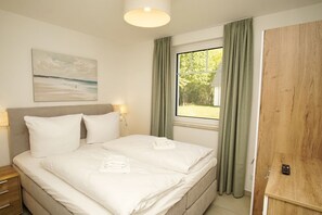 Un dormitorio con cama, una mesita de noche, lámpara, un cuadro, una ventana con cortinas y una puerta de madera.