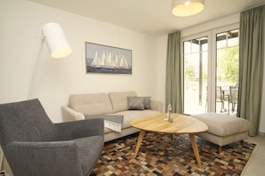 A living room with a sofa, a chair, a coffee table, and a floor lamp. There is a window with curtains and a painting on the wall.
