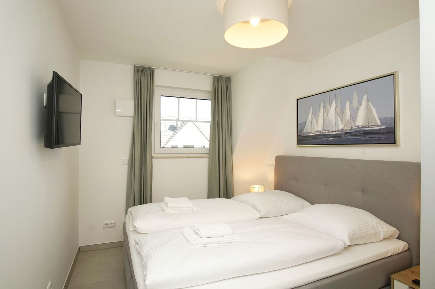 Une chambre d’hôtel avec un lit, un téléviseur fixé au mur, une fenêtre avec des rideaux et un tableau représentant des voiliers.