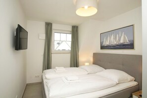 A hotel room with a bed, a TV mounted on the wall, a window with curtains, and a painting of sailboats.