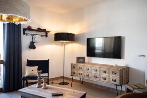 A modern living room with a flat-screen TV mounted on the wall, a wooden cabinet with drawers, a black chair, a floor lamp, and a table with a candle.