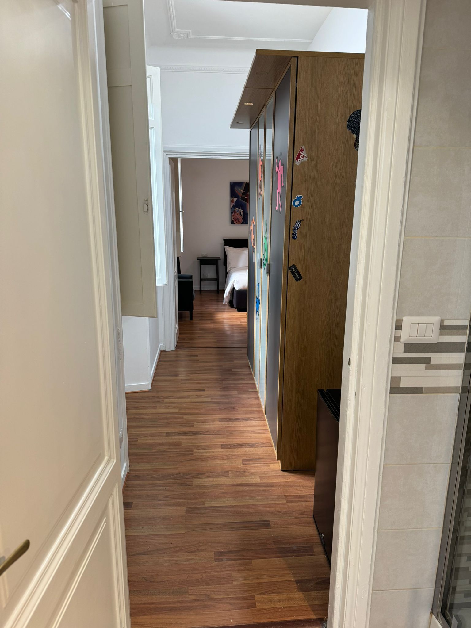 A hallway with wooden flooring leading to a bedroom with a bed and a nightstand.
