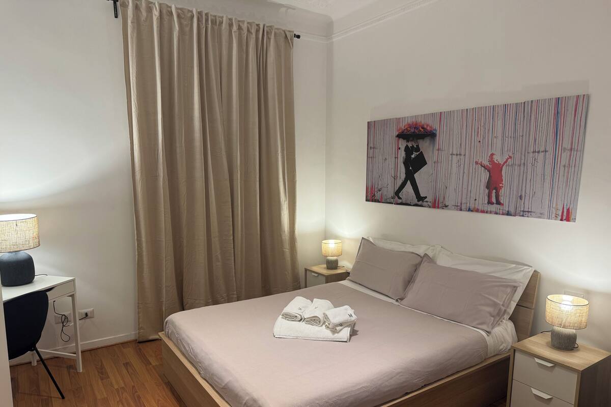 Hotellihuone, jossa on sänky, työpöytä, tuoli, yöpöytä ja taulu seinällä.