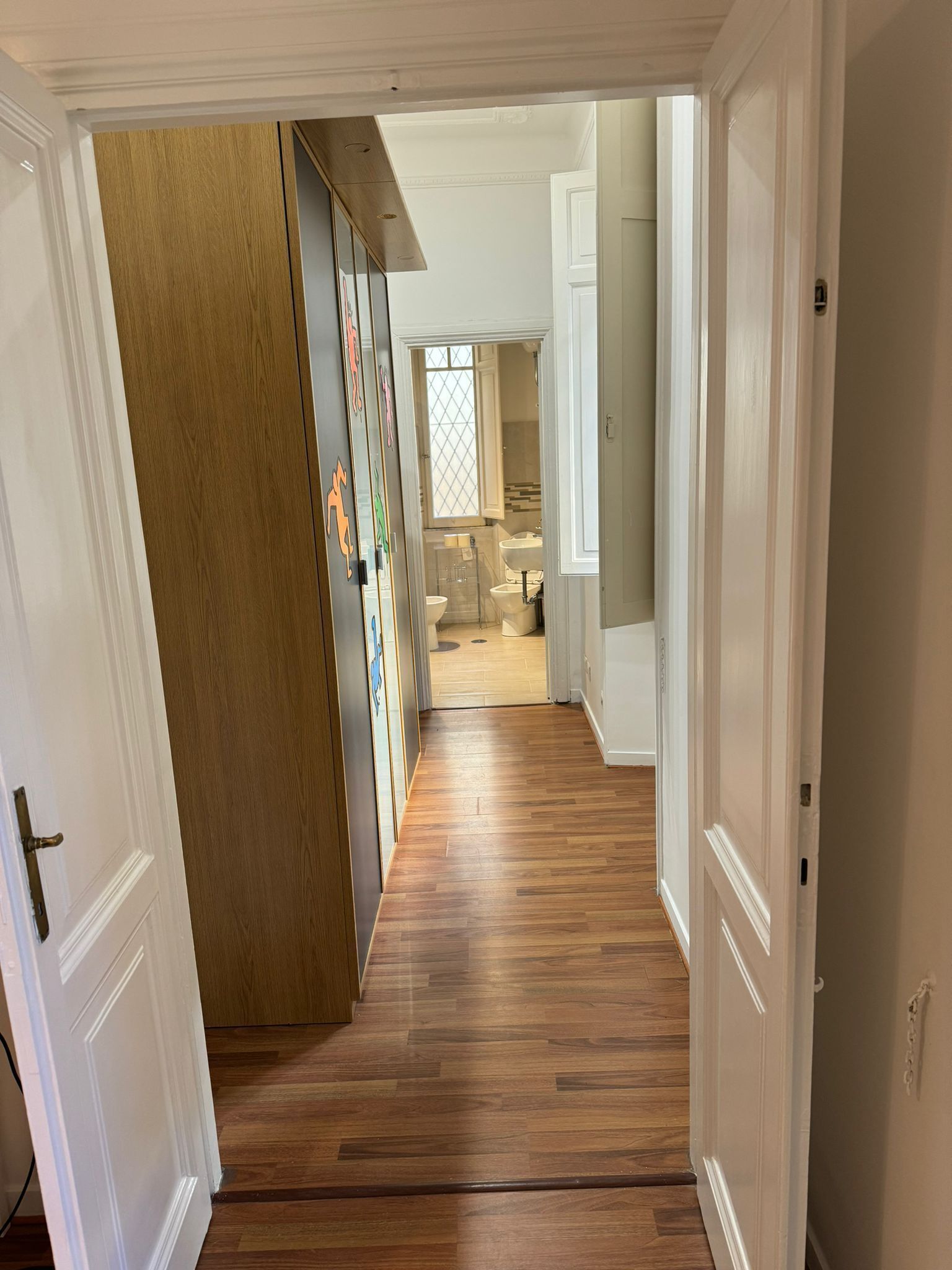 Un couloir au plancher de bois menant à une salle de bain dotée d’une fenêtre et d’un lavabo.