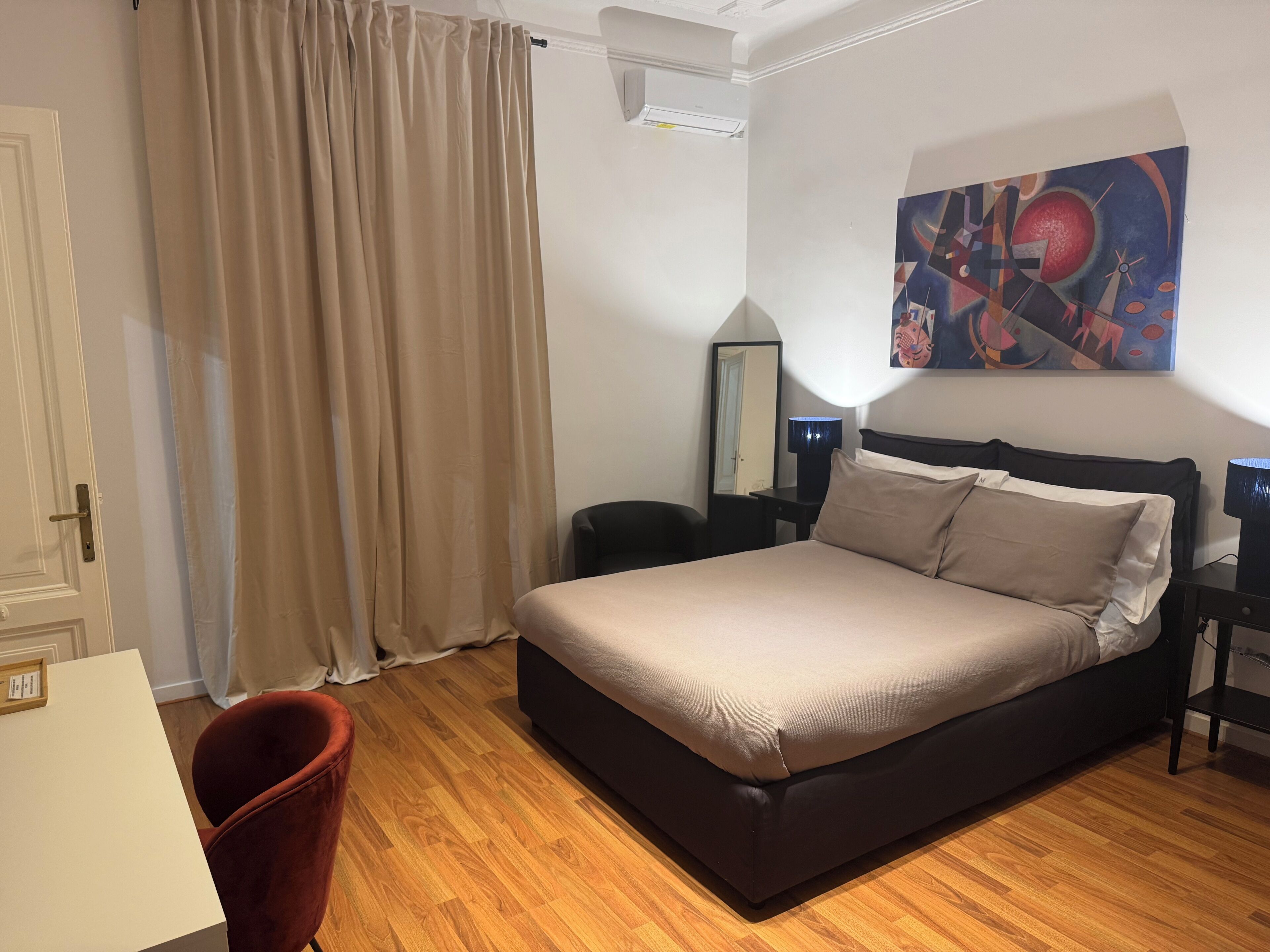 A hotel room with a bed, a desk, a chair, a mirror, and a painting on the wall.