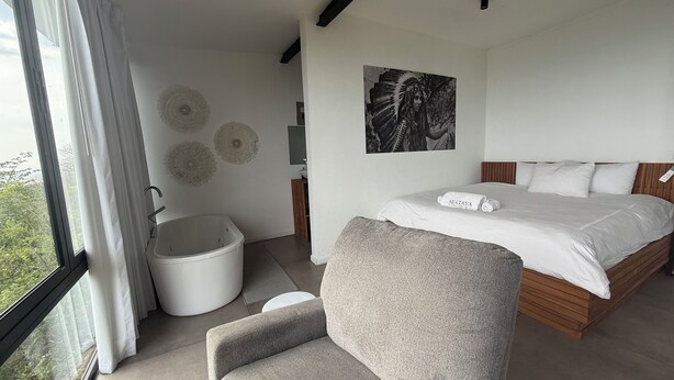 A modern bedroom with a large bed, a wooden headboard, a gray armchair, a white bucket, and a wall-mounted toilet.