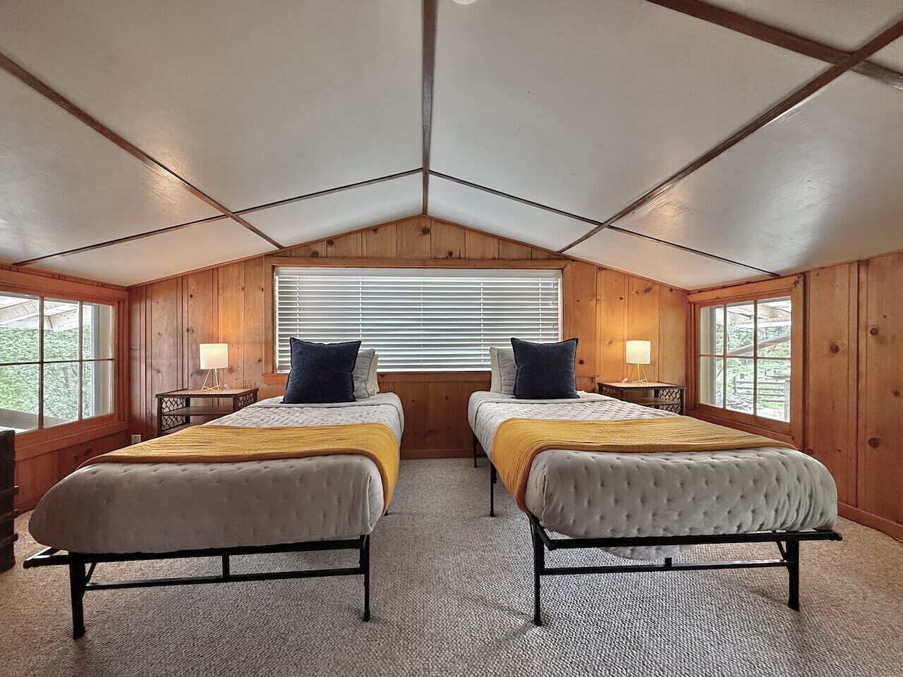 A room with two beds, wooden walls, a sloped ceiling, and large windows.