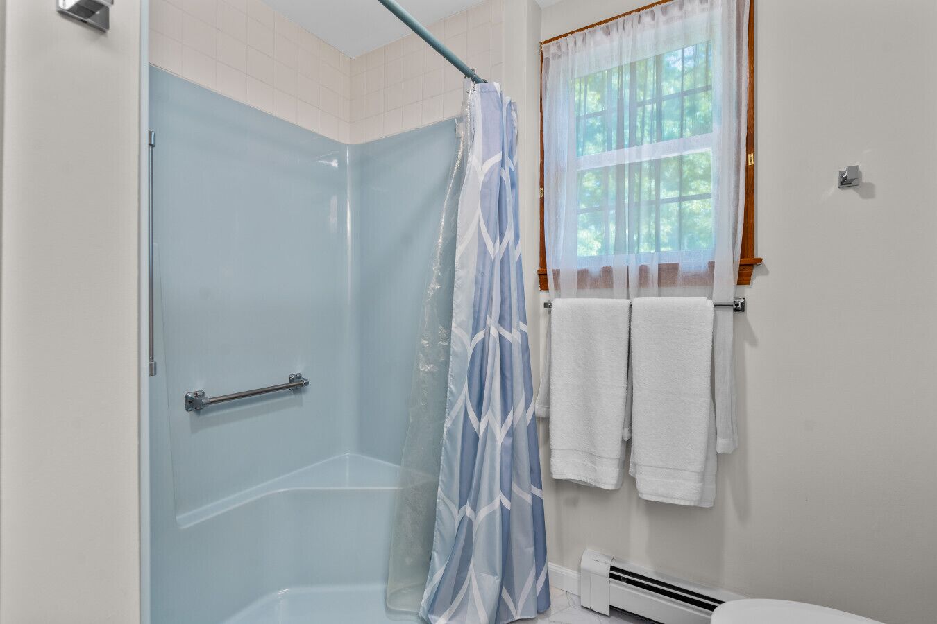 A bathroom with a glass shower enclosure, a towel rack with white towels, and a window with a sheer curtain.