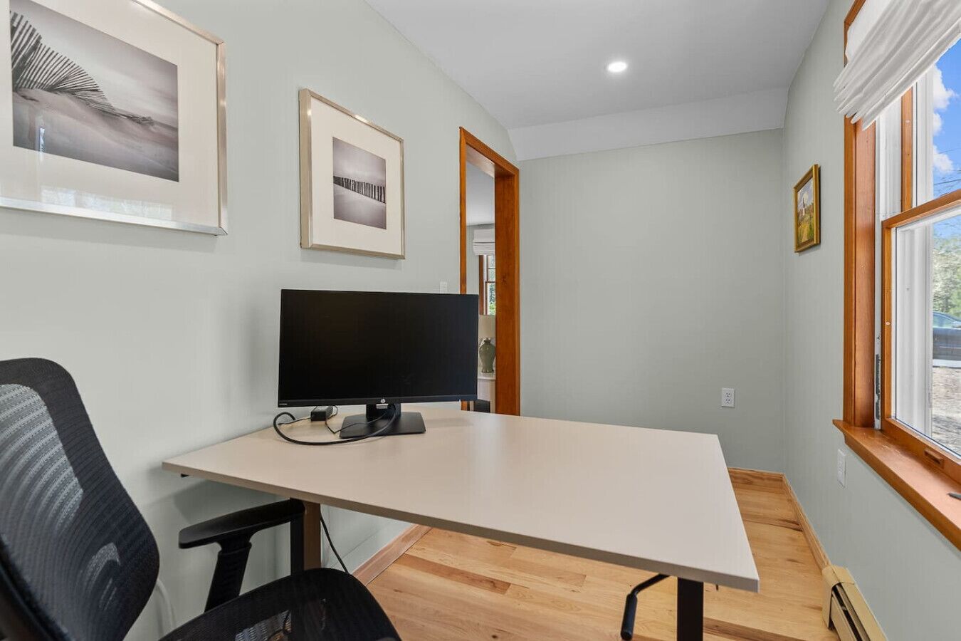 A modern office space with a desk, computer, and chair. There are framed pictures on the wall and a window with a view.
