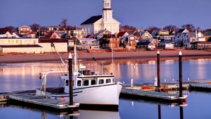 A harbor with a white boat docked at a pier, a lighthouse in the background, and a row of houses along the shore.