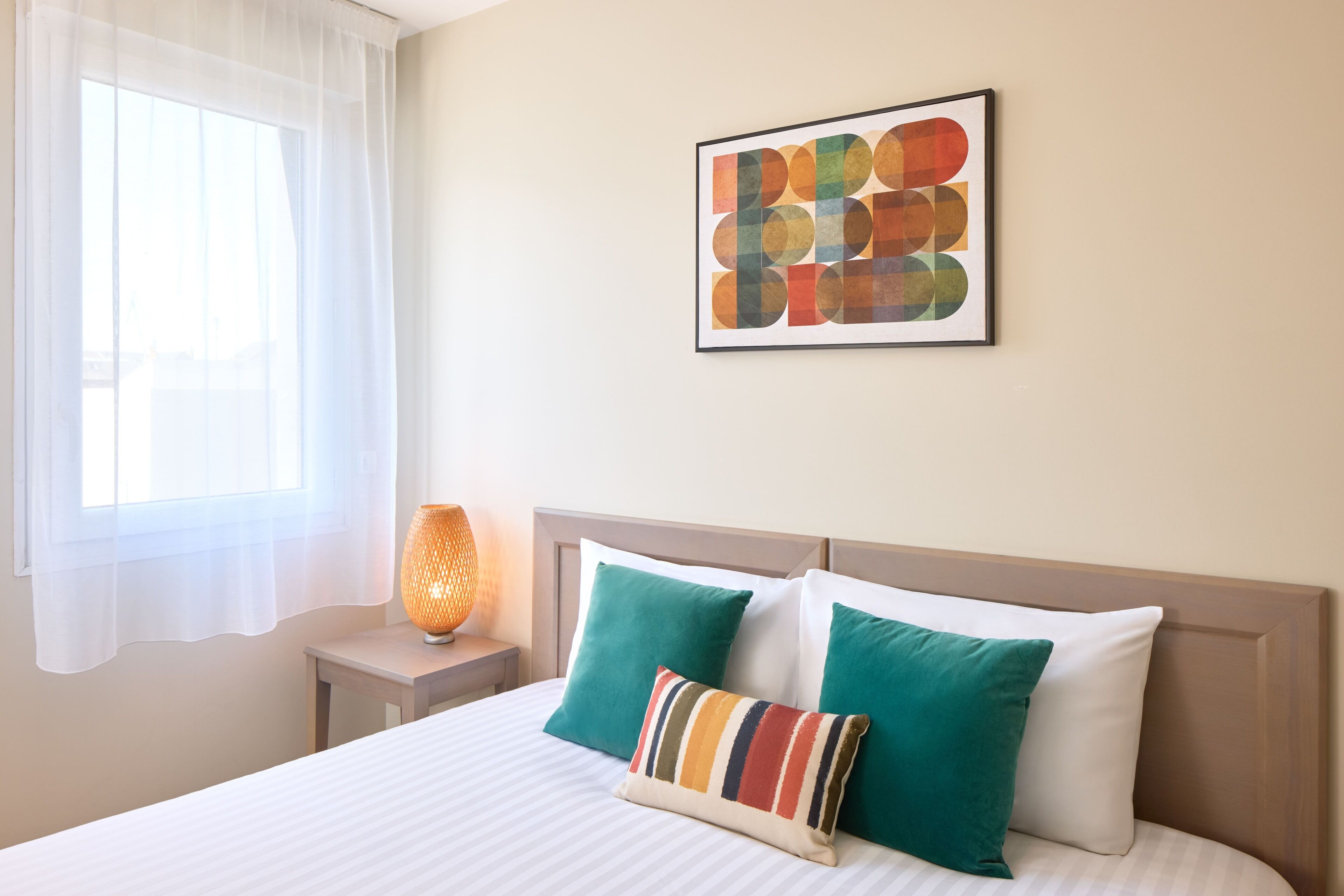 A hotel room with a bed, pillows, a nightstand, a lamp, and a window with curtains.