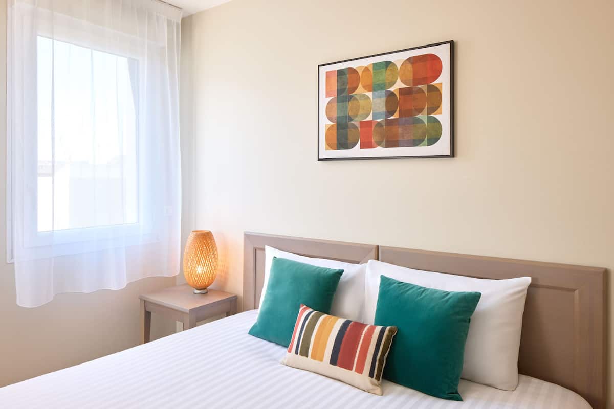 Une chambre d’hôtel avec un lit, des oreillers, une table de chevet, une lampe et une fenêtre avec des rideaux.