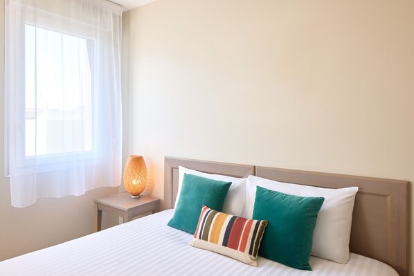 Une chambre d’hôtel avec un lit, deux oreillers, une lampe de chevet et une fenêtre avec des rideaux légers.