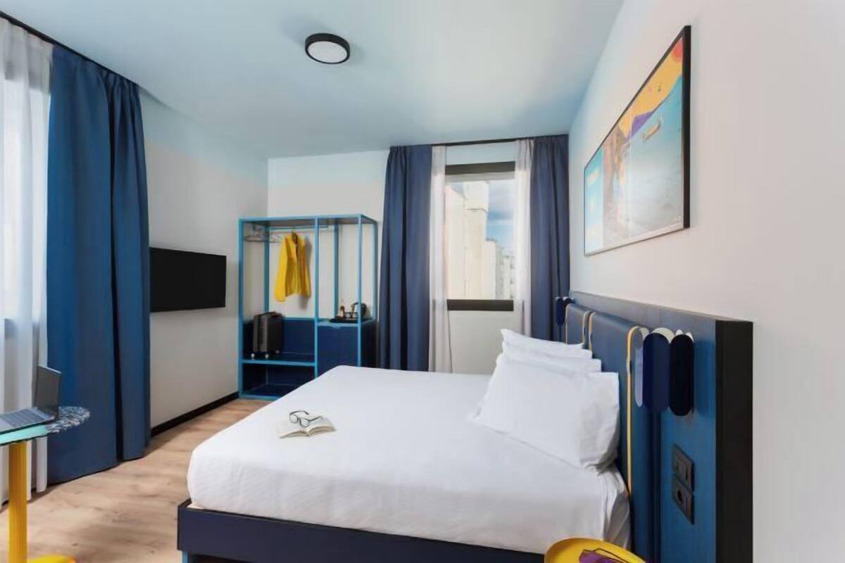 Ett hotellrum med en säng, ett skrivbord med en bärbar dator, en klädställning och en tavla på väggen.